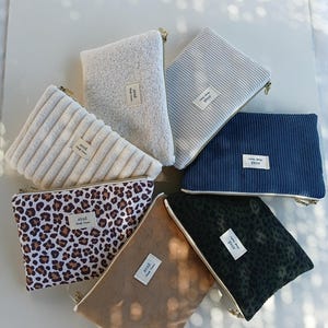 Puede incluir: Una colección de neceseres con cremallera en varios colores y texturas, incluyendo pana, sherpa e impresión de leopardo. Cada neceser tiene una pequeña etiqueta rectangular con el texto "AVAE" y una cremallera dorada.