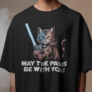 以下が含まれることがあります： 青いライトセーバーを持った漫画の猫が描かれた黒いTシャツ。猫はフード付きのローブを着ています。下の白い文字は「MAY THE PAWS BE WITH YOU」です。