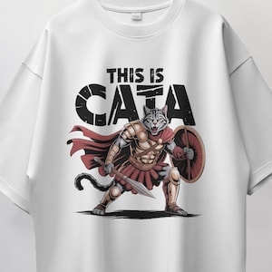May include: White t-shirt featuring a cartoon illustration of a cat warrior with armor, a spear, and a shield. The text "THIS IS CATA" is printed above the cat. The cat is in a battle stance, with a red cape.