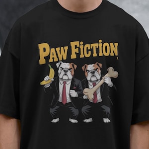 May include: Black t-shirt featuring the text "Paw Fiction" in gold lettering. The graphic depicts two bulldog characters in suits, one holding a banana and the other a bone. The shirt is worn by a person.