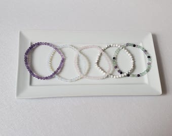May include: Five beaded bracelets in shades of purple, white, pink, and green are arranged on a white rectangular plate. The bracelets appear to be made of small, round gemstones. The background is a plain white surface.