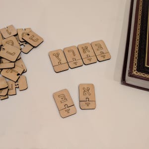 Puede incluir: Piezas de rompecabezas de madera con letras hebreas dispersas sobre una superficie blanca. Las piezas son de color marrón claro con letras grabadas en marrón oscuro. Un libro con una cubierta marrón y ribete dorado está en el fondo.