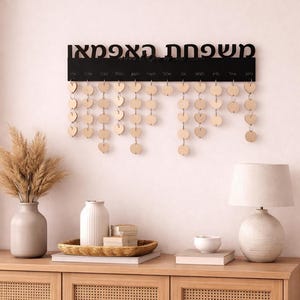 May include: A black wooden family calendar with Hebrew text at the top and wooden heart-shaped tags hanging below. The calendar is mounted on a light pink wall above a wooden cabinet with decorative items.