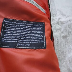 May include: A bright orange interior lining of a jacket featuring a black rectangular care instruction label with white text that reads CARE INSTRUCTIONS FOR YOUR AUTHENTIC AVIREX JACKET. The jacket has a white leather exterior and a metal zipper.