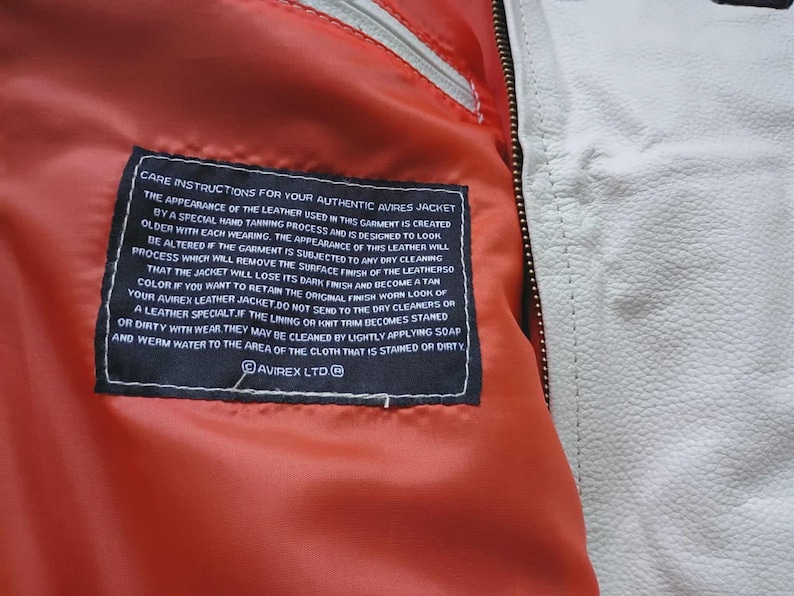 May include: A bright orange interior lining of a jacket featuring a black rectangular care instruction label with white text that reads CARE INSTRUCTIONS FOR YOUR AUTHENTIC AVIREX JACKET. The jacket has a white leather exterior and a metal zipper.