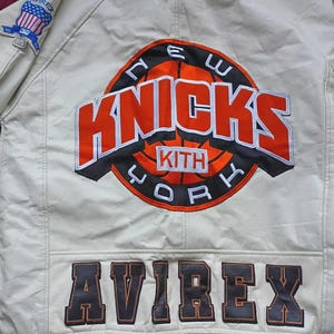 May include: An off-white leather jacket featuring a large embroidered New York Knicks basketball logo on the back with the text KITH in the center. The word AVIREX is stitched in large dark letters at the bottom, and an Avirex flag patch is on the sleeve.