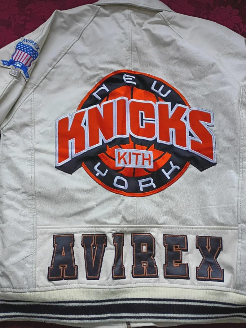 May include: An off-white leather jacket featuring a large embroidered New York Knicks basketball logo on the back with the text KITH in the center. The word AVIREX is stitched in large dark letters at the bottom, and an Avirex flag patch is on the sleeve.