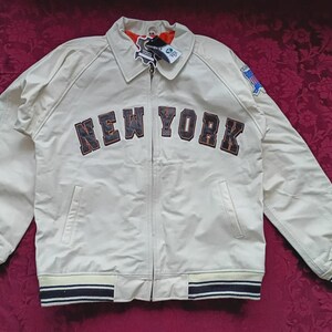 May include: A cream-colored zip-up jacket featuring the text NEW YORK in dark lettering across the chest. The jacket includes a collar, long sleeves with striped ribbed cuffs, a matching striped ribbed hem, and a small blue patch on the left sleeve.