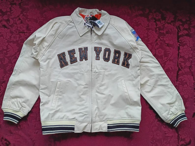 May include: A cream-colored zip-up jacket featuring the text NEW YORK in dark lettering across the chest. The jacket includes a collar, long sleeves with striped ribbed cuffs, a matching striped ribbed hem, and a small blue patch on the left sleeve.
