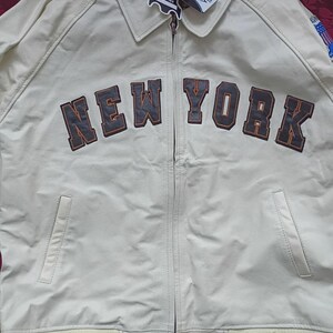 May include: An off-white leather jacket featuring a full-length front zipper, a classic collar, and two side pockets. The chest displays the text NEW YORK in dark brown block letters with orange outlines. It includes a ribbed hem and cuffs.