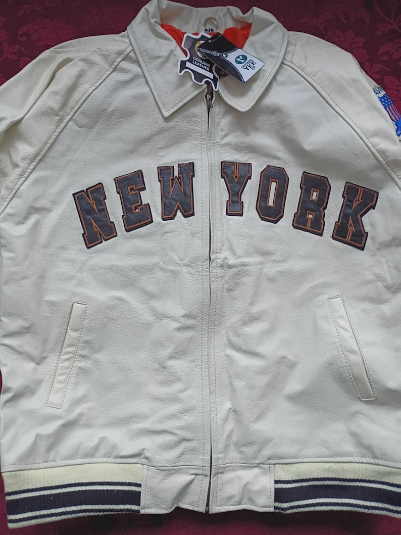 May include: An off-white leather jacket featuring a full-length front zipper, a classic collar, and two side pockets. The chest displays the text NEW YORK in dark brown block letters with orange outlines. It includes a ribbed hem and cuffs.