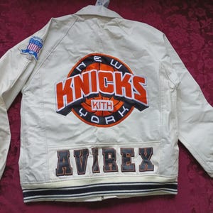 May include: An off-white jacket featuring a large embroidered New York Knicks and Kith logo on the back. The word AVIREX is stitched in dark lettering at the bottom. A small American flag patch is on the right sleeve, and the cuffs have stripes.