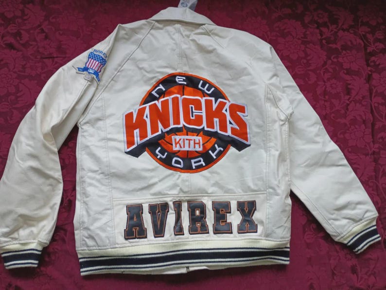May include: An off-white jacket featuring a large embroidered New York Knicks and Kith logo on the back. The word AVIREX is stitched in dark lettering at the bottom. A small American flag patch is on the right sleeve, and the cuffs have stripes.