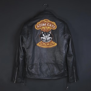 Op de afbeelding: Zwarte leren jas met ritssluiting en een decoratieve patch op de rug. De patch toont een cartoonkat in een paddenstoelwolk met de tekst "ATOM CATS" in een cirkel.
