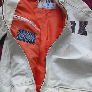May include: A white leather jacket with a bright orange interior lining. The jacket features a front zipper closure, a ribbed hem with black and white stripes, and a dark blue patch with the text RK on the front. A tag reads Avirex USA New York.