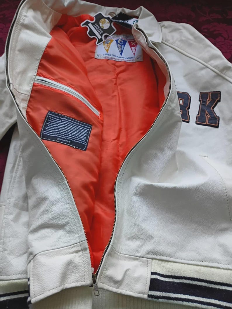 May include: A white leather jacket with a bright orange interior lining. The jacket features a front zipper closure, a ribbed hem with black and white stripes, and a dark blue patch with the text RK on the front. A tag reads Avirex USA New York.