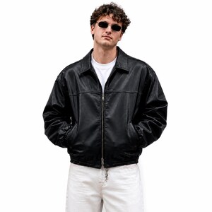 May include: A black leather jacket with a classic collar, a full-length silver metal zipper, and ribbed cuffs. The jacket is worn over a plain white t-shirt, paired with white denim jeans, and accessorized with dark-tinted rectangular sunglasses.