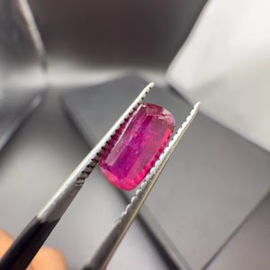 May include: A vibrant, rectangular-cut ruby gemstone, held by tweezers. The ruby displays a deep, saturated pink-red hue, with visible facets. The tweezers are silver-colored, and the background is a dark gray.