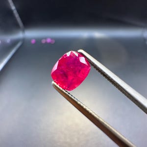 May include: A vibrant, cushion-cut ruby gemstone held by silver tweezers. The ruby displays a deep red hue and a faceted surface, reflecting light. The background is a dark, blurred gradient, highlighting the gemstone's brilliance.