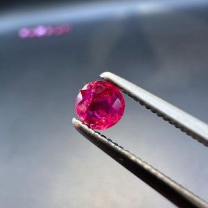 Könnte beinhalten: Ein facettierter, runder, tiefroter Edelstein wird von einer silbernen Pinzette vor einem dunklen Hintergrund gehalten. Der Edelstein hat eine lebendige, gesättigte Farbe mit sichtbaren inneren Strukturen. Die Pinzette ist scharf fokussiert und hebt die Klarheit des Edelsteins hervor.