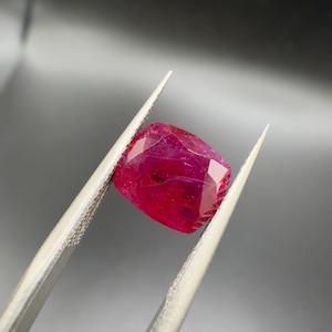 Könnte beinhalten: Ein facettierter, kissenförmiger Rubin, der von einer Pinzette gehalten wird. Der Rubin hat eine tiefe, gesättigte rote Farbe mit sichtbaren inneren Einschlüssen. Die Pinzette ist silberfarben und reflektiert das Licht.