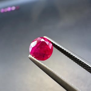 May include: A faceted, round-cut ruby gemstone held by silver tweezers. The ruby is a deep, vibrant red color, with visible internal inclusions. The background is a blurred, dark gray.