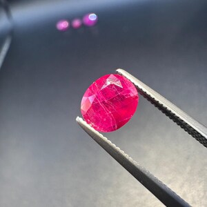 May include: A faceted, oval-shaped ruby gemstone held by tweezers. The ruby is a vibrant, deep red color with internal inclusions visible. The background is dark, highlighting the gem's brilliance.
