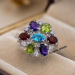 May include: A silver-toned ring featuring a central oval blue gemstone surrounded by a cluster of oval-cut purple, green, and dark red gemstones, accented with small clear stones. The text Chayna Jewels is printed on the side of the ring.