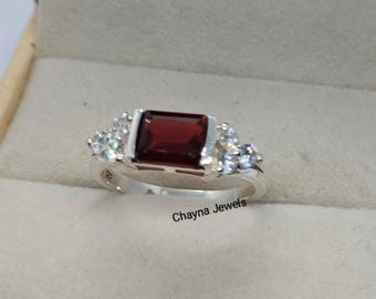 May include: A silver ring with a rectangular, deep red gemstone as the centerpiece. The ring is flanked by clusters of smaller, clear gemstones. The text "Chayna Jewels" is visible on the ring.