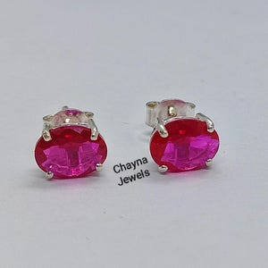 Puede incluir: Un par de pendientes de botón ovalados con piedras rosas facetadas engastadas en garras de metal plateado. El texto Chayna Jewels está impreso en negro entre los dos pendientes, que se muestran sobre un fondo liso y claro.