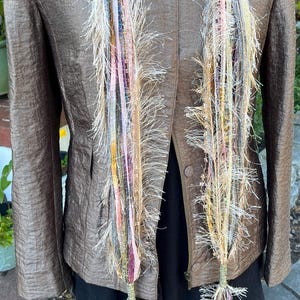 May include: A brown jacket with a textured finish is paired with a colorful, fringed scarf. The scarf features strands of various colors, including pink, yellow, and blue, creating a unique and artistic accessory. The jacket has a classic collar and button closure.