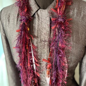May include: A vibrant, handmade scarf with a textured, fringed design. The scarf features a mix of deep red, purple, and orange hues, with accents of white and yellow. The scarf is draped over a gray collared shirt.