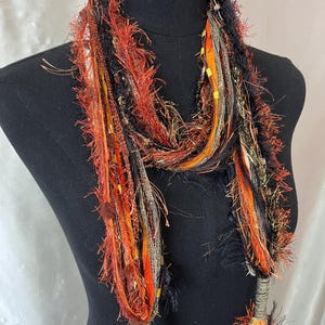 May include: A multi-textured scarf featuring a blend of black, orange, and metallic gold yarns. The design incorporates fringed, fuzzy, and ribbon-like strands woven together, with braided sections at the ends that terminate in decorative tassels.