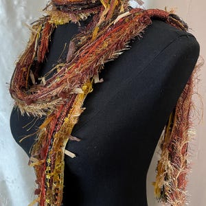 May include: A multi-textured scarf featuring a blend of yarn and ribbon in shades of rust, gold, brown, and copper. The scarf is composed of various strands, including fuzzy, metallic, and flat ribbon textures, draped around a black mannequin.