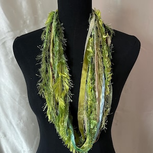 May include: A green and gold infinity scarf with a variety of textures, including fringe and ribbons, displayed on a black mannequin. The scarf features shades of green, gold, and a hint of blue.
