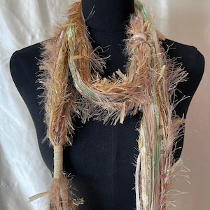 May include: A decorative scarf with a textured, fringed design. The scarf features a mix of gold, tan, and pastel colors, with a variety of ribbons and threads. The scarf is displayed on a black mannequin, showcasing its draping effect.