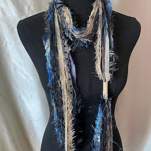May include: A multi-textured scarf featuring a blend of blue, black, and metallic gold fibers. The design incorporates various yarn types, including fringed, ribbon-like, and smooth strands, creating a layered, decorative appearance around the neck.