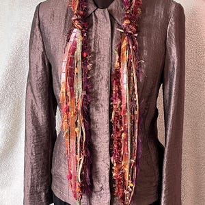 May include: A colorful, handmade scarf with a textured, fringed design. The scarf features a mix of red, orange, purple, and gold threads, creating a vibrant and eye-catching accessory. The scarf is draped over a brown blazer.