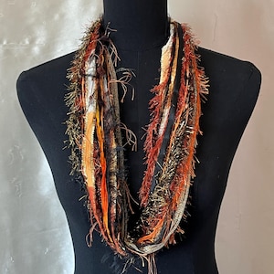 May include: A multi-textured infinity scarf featuring a blend of orange, black, gold, and metallic copper fibers. The scarf is composed of various ribbon and yarn strands, including fringed, shimmering, and smooth textures, creating a layered look.