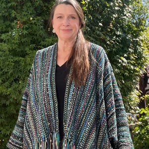 May include: A multi-colored woven shawl with fringe. The shawl features a mix of teal, black, and pink threads. The fringe at the bottom has gold and teal ribbons. The person in the image is wearing a black top.