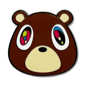 Puede incluir: Un pin con la cara de un oso marrón con un contorno negro. El oso tiene ojos grandes y coloridos con acentos rosas, azules y amarillos. Las orejas y el hocico del oso son de color beige claro.
