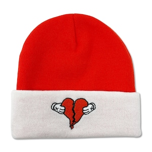 Puede incluir: Un gorro rojo con un puño blanco. El puño presenta un diseño bordado de un corazón roto con alas. El gorro está hecho de un material de punto suave, perfecto para el clima frío.