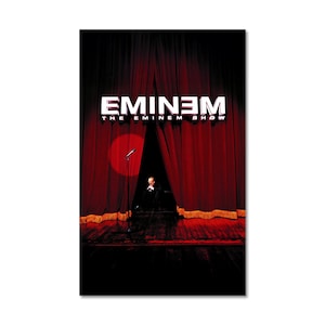May include: A poster featuring a stage with red curtains, a microphone, and a spotlight. The text "EMINEM" is displayed in white block letters above the words "THE EMINEM SHOW". A figure is visible on the stage.