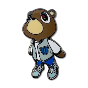 Puede incluir: Un pin de esmalte de oso marrón que lleva una chaqueta universitaria azul claro y blanca con una "U" azul en la parte delantera. El oso también lleva vaqueros azules y zapatillas de deporte azul claro. El pin tiene un borde plateado.