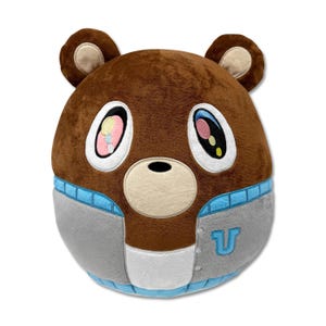 Puede incluir: Oso de peluche marrón con ojos grandes y expresivos y orejas pequeñas. El oso lleva una chaqueta gris y azul con una "U" en el pecho. Los ojos del oso tienen detalles en rosa, amarillo y azul. Un juguete suave y tierno.