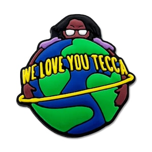 May include: A colorful shoe charm featuring a cartoon figure with black hair and glasses, hugging a globe. The globe is blue and green with a yellow band around it. The text "WE LOVE YOU TECCA" is in yellow.