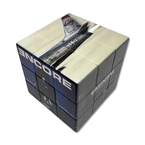 May include: A cube puzzle featuring an image of a silver airplane with the text "FU-20" and "TKCJS". The word "ENCORE" is printed in white on one side, and "EMINEM" on another.