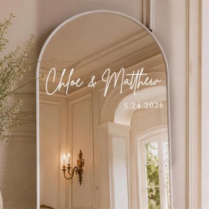 Op de afbeelding: Een gebogen spiegel met een wit frame. De spiegel reflecteert een kamer met een kroonluchter en een raam. De namen "Chloe & Matthew" en de datum "5.24.2026" staan in witte letters op de spiegel. De spiegel is een decoratief item.
