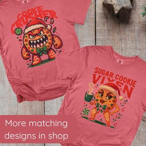 May include: Two red t-shirts with Christmas cookie designs. One shirt features a cookie monster with the text "Cookie Crusher." The other shirt has a cookie character with the text "Sugar Cookie Vixen." The shirts are on a wooden surface.