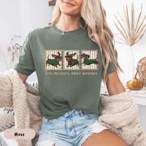 May include: Moss green t-shirt featuring three dachshunds wearing sweaters and glasses, each framed in a striped border. The text "less meanies, more weenies" is printed below the dogs. The shirt is made of a soft material.